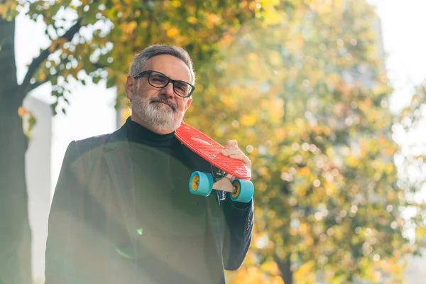 Active leisure time. Skating as sport for everyone. Elegant caucasian gray-haired businessman holding small neon-orange skateboard on his arm. Sunshine in camera. Autumn time. High quality photo
