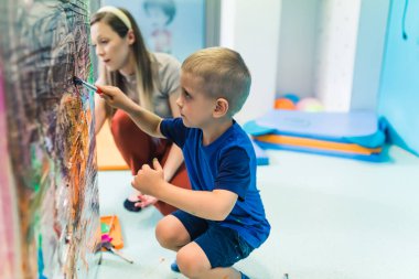 Mavi elbiseli, elinde fırça tutan, kreşteki selofana resim yapan, arka planda öğretmenlik yapan beyaz bir çocuk. Yüksek kalite fotoğraf