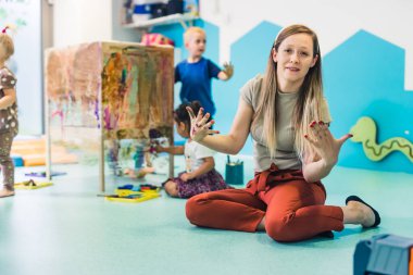 Beyaz bir öğretmen yerde boyalı elleriyle oturuyor ve kameraya bakıyor. Çocuklar arka planda elleriyle resim yapıyor. Anaokulu konsepti. Yüksek kalite fotoğraf