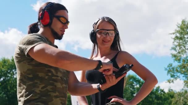 屋外射撃場 プロの射撃インストラクター 保護眼鏡の中年の筋肉ひげを生やした男性 若い女性のクライアントに基本的な銃の使い方を教えています 美しい夏の天気 — ストック動画