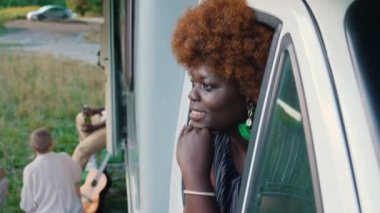 Dost canlısı Afro-Amerikalı kadın çenesine dokunuyor, gülümsüyor ve karavan penceresinden dışarı bakıyor. Yaz tatili dinlenme ve rahatlama zamanı. Yüksek kalite 4k görüntü