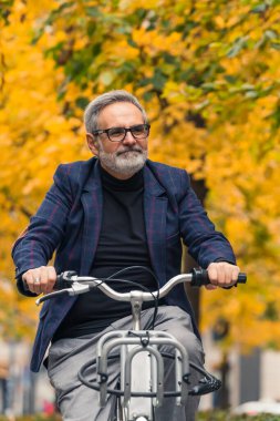 Şık ve mutlu sakallı gri saçlı olgun bir adam sonbahar mevsiminde ağaçların etrafında sarı yapraklarla bisiklet sürerken sıcak havanın tadını çıkarıyor. Yüksek kalite fotoğraf