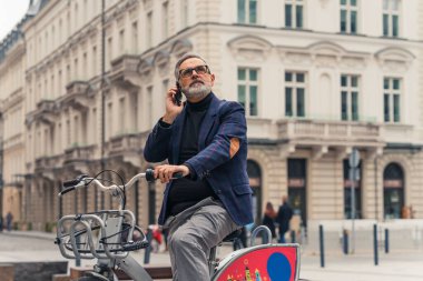 Sakallı, gri saçlı, olgun bir adam şehir sokaklarında bisiklet sürerken cep telefonuyla konuşuyor. Yüksek kalite fotoğraf
