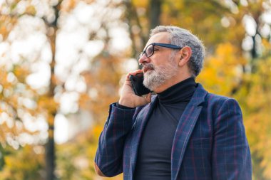 Yakışıklı, sakallı, gri saçlı, takım elbiseli olgun bir adam arkadaşıyla telefonda konuşuyor. Parkta yapraklar döküldü. Yüksek kalite fotoğraf