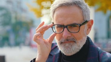 Gri sakallı, yakışıklı, yaşlı, gri saçlı bir adam yeni gözlüklerini tek eliyle düzeltiyor ve tek kaşını kaldırıyor. Arka plan bulanık. Açık hava çekimine yakın çekim. Yüksek kalite 4k görüntü