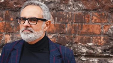 Hayat dolu Avrupalı, gri sakallı, iş kıyafetleri içinde kameraya yavaşça bakan beyaz bir adam. Arka plan olarak eski tuğla duvar. Göz giyimi konsepti. Yüksek kalite 4k görüntü