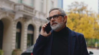 Açık hava orta boy yakın çekim. Yakışıklı, orta yaşlı, beyaz sakallı iş adamı şehirde dolaşıyor ve modern akıllı telefonuyla biriyle sohbet ediyor. Yavaş çekim klibi. Yüksek
