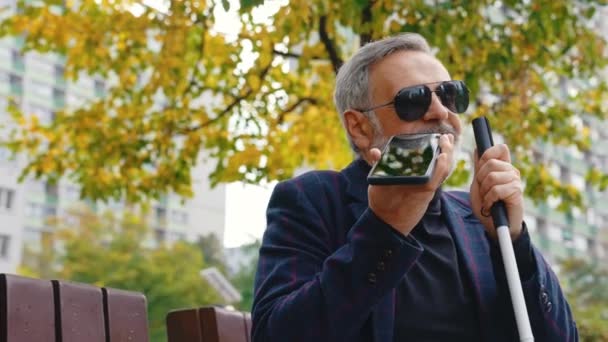 年长的视力残疾的高加索人手持步行起重机 戴着太阳镜 在他最喜爱的公园享受新鲜空气 并在电话中通过扩音器交谈 室外特写镜头 — 图库视频影像