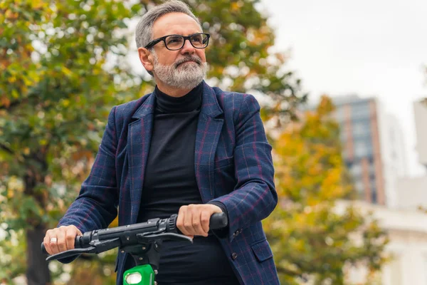 Millennial happy bearded grey-haired mature man with eyeglasses electro scooter riding and enjoying warm weather during the fall season. High quality photo