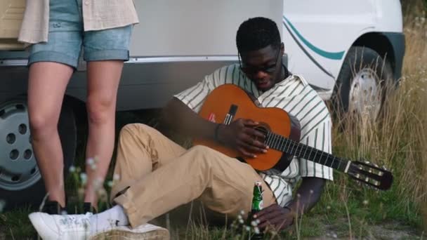 キャンパーバンのギターとビールボトルを持って地面に座ってサングラスの若い大人の黒人男性 友人のグループのキャンプ 水平屋外ビデオ 高品質4K映像 — ストック動画