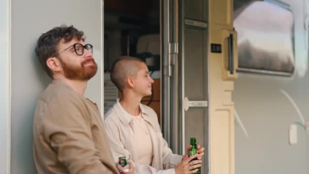两个年轻的成年朋友抱着啤酒瓶笑着坐在野营车外环顾四周 横向室外视频 高质量的4K镜头 — 图库视频影像