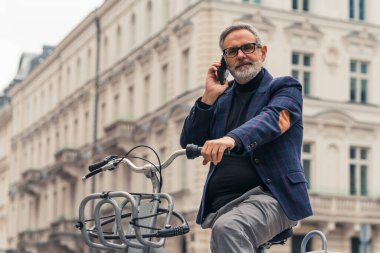 Sakallı, gri saçlı, olgun bir adam şehrin sokaklarında güzel binalarla bisiklet sürerken cep telefonuyla konuşuyor. Yüksek kalite fotoğraf