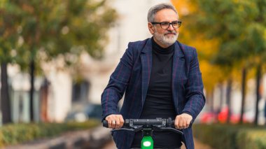 Milenyum mutlu, sakallı, gözlüklü, gri saçlı, olgun bir adam. Parkta elektro scooter sürüyor ve sonbahar sezonunda sıcak havanın tadını çıkarıyor. Yüksek kalite fotoğraf