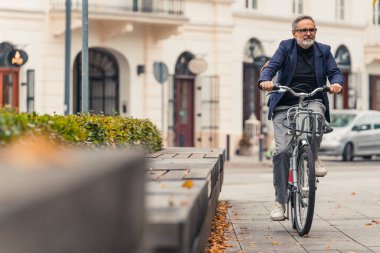 Bin yıllık mutlu sakallı, gri saçlı, gözlüklü olgun bir adam sonbahar mevsiminde şehrin sokaklarında bisiklet sürerken sıcak havanın tadını çıkarıyor. Yüksek kalite fotoğraf