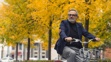 Şık ve mutlu sakallı gri saçlı olgun bir adam sonbahar mevsiminde ağaçların etrafında sarı yapraklarla bisiklet sürerken sıcak havanın tadını çıkarıyor. Yüksek kalite fotoğraf
