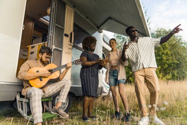 Farklı etnik kökenli modern gezginler güzel havanın tadını çıkarıyorlar, minibüslerinin dışında vakit geçiriyorlar, canlı gitar müziği eşliğinde dans ediyorlar. Karavan konsepti. - Evet. Yüksek kalite fotoğraf