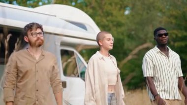 Bir grup tuhaf insan güzel bir çayırın ortasında duruyor ve mutlu bir şekilde havaya zıplıyorlar. Arkalarında beyaz bir karavan var. Yaz seyahati. Yüksek kalite 4k görüntü