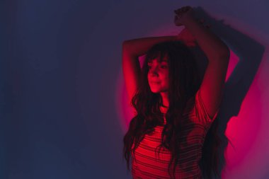 Güzel, sıradan giyinmiş, uzun saçlı, beyaz bir kız. Kolları havada poz veriyor, mor duvara yaslanıyor ve kırmızı daire ışığına bakıyor. Modern estetik konsept. Yüksek kalite fotoğraf