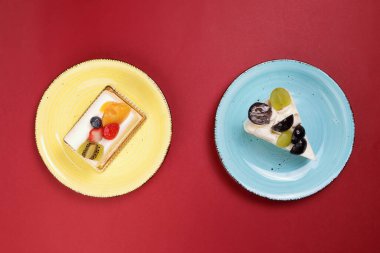 Kırmızı zemin üzerinde yeşil mavi yuvarlak tabakta iki dilim yeşil üzüm Kivi çilekli mango meyveli kremalı pasta.