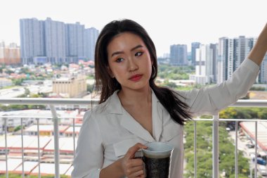 Güneydoğu Asyalı güzel bir kadın elinde çay fincanı tutuyor. Balkon pencere manzarası arka planında. Rahatlatıcı bir düşünce.