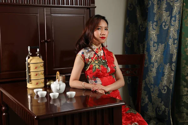 魅力的な若いです女性身に着けていますレトロな中国のChongsamドレス座って隣にヴィンテージテーブルとともに茶セットティフィンキャリアチェア探して保持チェックポーズ — ストック写真