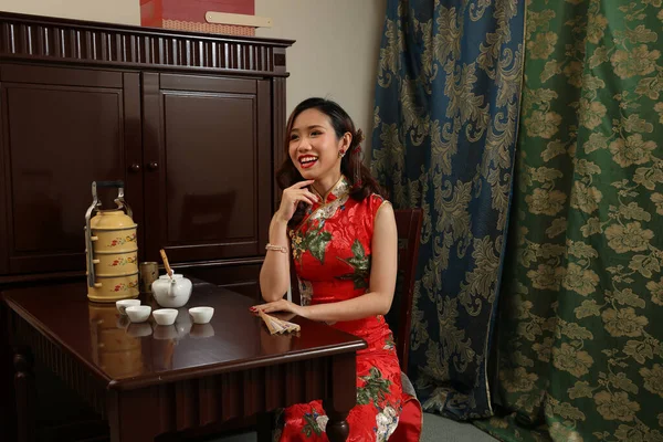 魅力的な若いです女性身に着けていますレトロな中国のChongsamドレス座っている隣にヴィンテージテーブルとともに茶セットティフィンキャリアチェア探して保持チェックポジションポイント笑顔 — ストック写真