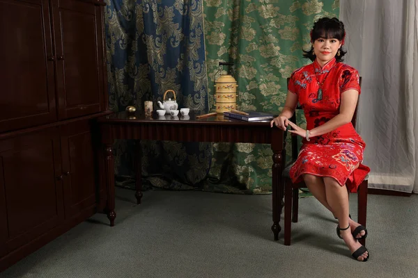 魅力的な若いです女性身に着けていますレトロな中国のChongsamドレス座って隣にヴィンテージテーブルとともに茶セットティフィンキャリアチェア探して保持チェックポーズ — ストック写真
