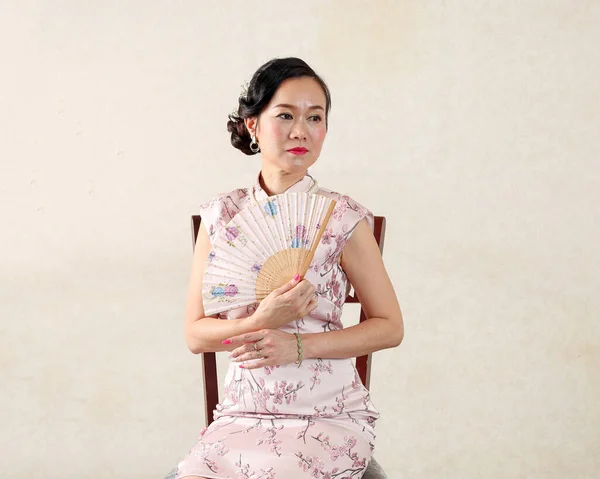 魅力的な中年女性身に着けている中国のChongsamドレス座って上の椅子保持中国の装飾された手のファン — ストック写真