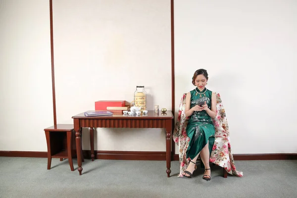現代的な中国スタイルのドレスを身に着けている美しい若い女性Chengsamファッションレトロヴィンテージテーブルルームスマートフォンを使用して — ストック写真