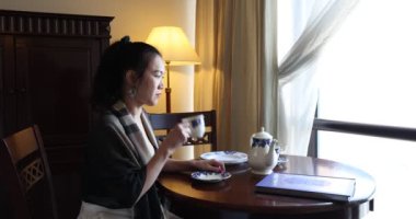 Asya kökenli, orta yaşlı çekici bir kadın retro-klasik masa sandalyesinde oturuyor. Dolap zeminindeki lamba seti, pencere ışığı çay içiyor. 