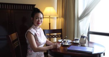 Asya kökenli, orta yaşlı, Cheongsam 'lı çekici bir kadın. Eski moda masa sandalyesinde oturuyor. Dolap lambası. 