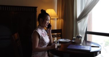 Asya kökenli, orta yaşlı, Cheongsam 'lı çekici bir kadın retro-klasik masa sandalyesinde oturuyor. 