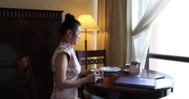 Asya kökenli, orta yaşlı, Cheongsam 'lı çekici bir kadın retro-klasik masa sandalyesinde oturuyor. 