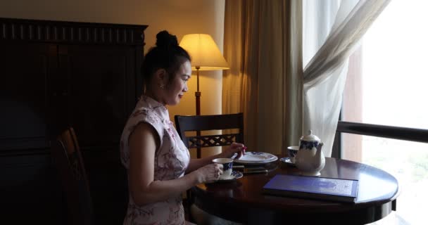 原产于亚洲的中年妇女 穿着旗袍坐在复古古时代的房间桌椅地板灯笼茶杯橱窗迪尔茶回音按摩托智能手机 — 图库视频影像