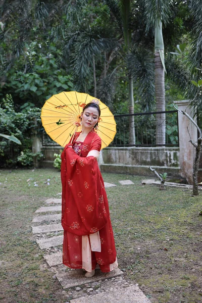 若い魅力的なアジアの女性は 伝統的な中国の赤い半布ロングスカートの衣装スカーフヘアピン耳飾り傘屋外緑の庭のパティオを身に着けている — ストック写真