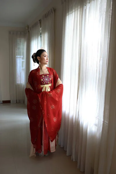 若い魅力的なアジアの女性を身に着けている伝統的な中国の赤い半布ロングスカートの衣装スカーフヘアピンピアス室内で窓の白いカーテンライト — ストック写真