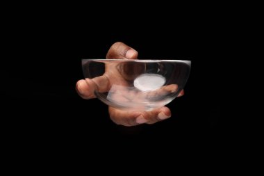 Asyalı, koyu tenli, tek elli, siyah arka planda küçük, şeffaf cam bir kase tutan parmak. 