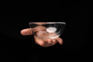 Asyalı, koyu tenli, tek elli, siyah arka planda küçük, şeffaf cam bir kase tutan parmak. 