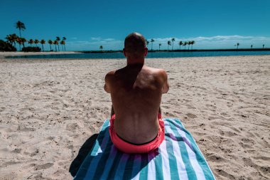 Turuncu şortlu çıplak gövdeli bir adam havluya oturup okyanusa bakar..