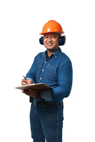 若いエンジニアはヘルメットとイヤーマフを着用し 長袖のシャツとズボンを着用しています クリップボードに立って メモを取り 喜んで笑顔 クリッピングパスで白い背景に隔離され — ストック写真