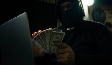 Hands of a man in a hoodie holding money and using a laptop digging into financial data-Internet theft concept