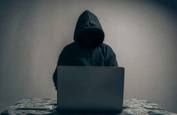 A hacker wears a long-sleeved shirt with a hood covering his head. He was sitting in a dark room using his laptop and had a lot of dollars on the table. Data theft concept. with copy space.