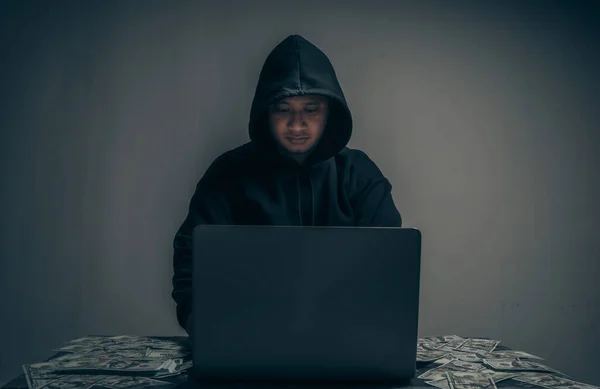 A hacker wears a long-sleeved shirt with a hood covering his head. He was sitting in a dark room using his laptop and had a lot of dollars on the table. Data theft concept. with copy space.