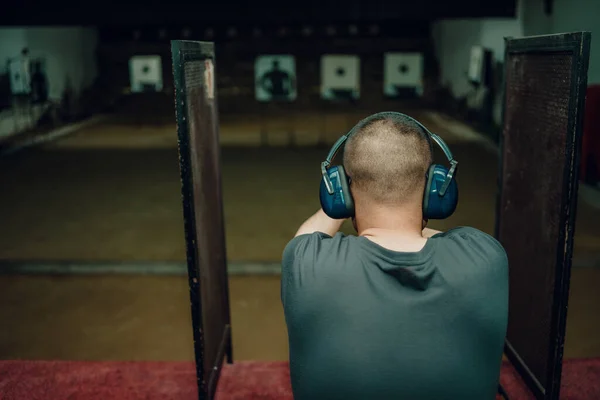 A professional man wearing earmuffs and goggles is practicing shooting a 9mm pistol inside a shooting range.