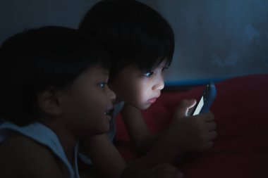 İki yakışıklı Asyalı çocuk karanlık bir yatak odasında cep telefonlarının üzerinde yatıyor. Smartphone Oynayan Bağımlı. Çocuklarda hiç korku yok..