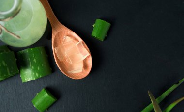 Ahşap bir kaşıkta Aloe Vera eti yanında dilimlenmiş aloe vera var ve cam bir şişede aloe vera jeli siyah bir taş yüzeyde duruyor..