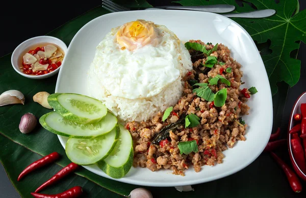 Thai street food, basil fried rice, minced pork, fried egg and sliced cucumber on a black table.