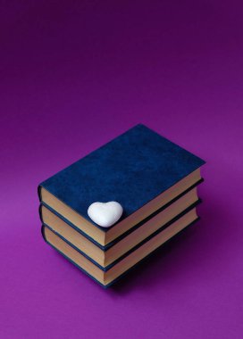 Kitaplar, kalın kapaklı, üst üste yığılmış, küçük beyaz bir kalp, mor bir arka planda. Metin uzayı. Kitap severler günü konsepti. Asgari biçim.