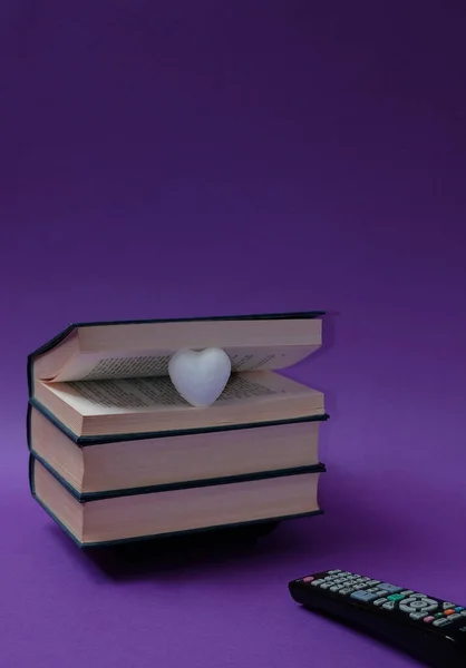 Books stacked on top of each other, a little white heart between the pages and a remote control on a purple background. Text space. Reading books vs. watching TV concept . Minimal style.