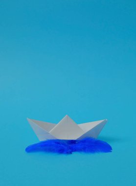 Mavi tüylü, gökyüzü mavisi arka planda beyaz kağıttan yapılmış bir tekne. Metin uzayı. Tatil zamanı, seyahat, yaratıcılık, konsept. Asgari biçim.